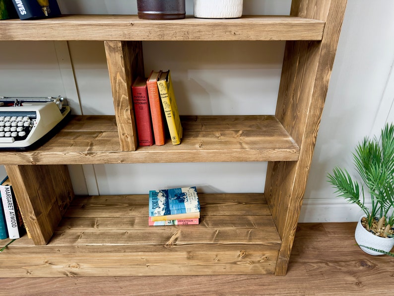 Puede incluir: Estanter&iacute;a r&uacute;stica de madera con tres estantes. Los estantes exhiben libros de varios colores, una m&aacute;quina de escribir vintage y objetos decorativos. La madera tiene un tono marr&oacute;n c&aacute;lido, con un dise&ntilde;o sencillo y funcional.