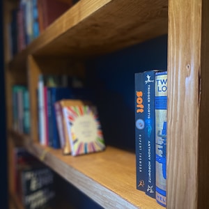 May include: A wooden bookshelf with two shelves, filled with books. The top shelf has a few books, while the bottom shelf is full of books. The books on the top shelf are out of focus, while the books on the bottom shelf are in focus. The books on the bottom shelf are a mix of different colors and sizes. The bookshelf is against a dark blue wall.