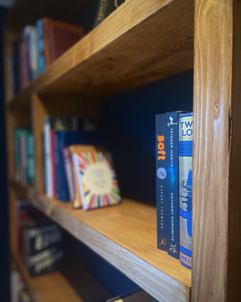 May include: A wooden bookshelf with two shelves, filled with books. The top shelf has a few books, while the bottom shelf is full of books. The books on the top shelf are out of focus, while the books on the bottom shelf are in focus. The books on the bottom shelf are a mix of different colors and sizes. The bookshelf is against a dark blue wall.