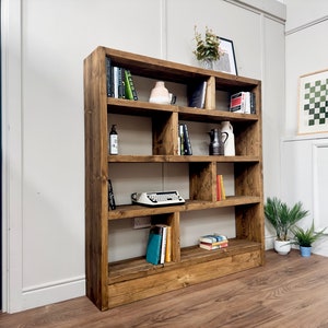 Puede incluir: Una gran estanter&iacute;a de madera r&uacute;stica con m&uacute;ltiples compartimentos. Los estantes est&aacute;n llenos de libros, objetos decorativos y una m&aacute;quina de escribir antigua. La madera tiene un acabado c&aacute;lido y natural, contra una pared blanca.