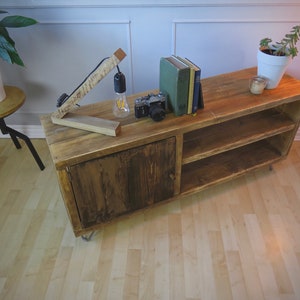 K&ouml;nnte beinhalten: Rustikaler TV-Schrank aus Holz mit Schrankt&uuml;r und offenen Regalen. Eine Vintage-Kamera, B&uuml;cher und eine kleine Lampe stehen obenauf. Der Schrank ist ca. 120 cm lang.