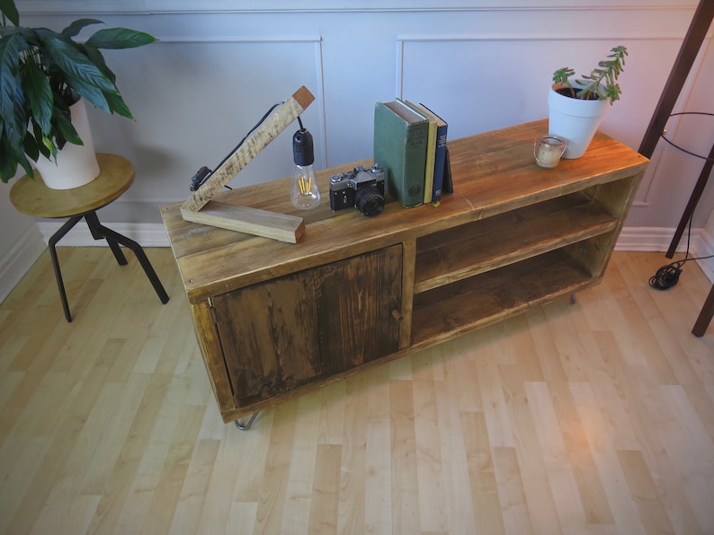 K&ouml;nnte beinhalten: Rustikaler TV-Schrank aus Holz mit Schrankt&uuml;r und offenen Regalen. Eine Vintage-Kamera, B&uuml;cher und eine kleine Lampe stehen obenauf. Der Schrank ist ca. 120 cm lang.