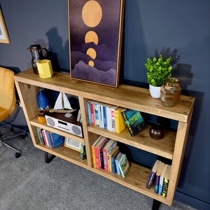 May include: A wooden bookshelf with black metal legs, filled with books, decorative items, and a framed artwork depicting golden orbs over a purple landscape. A small white radio and a miniature sailboat are also visible.