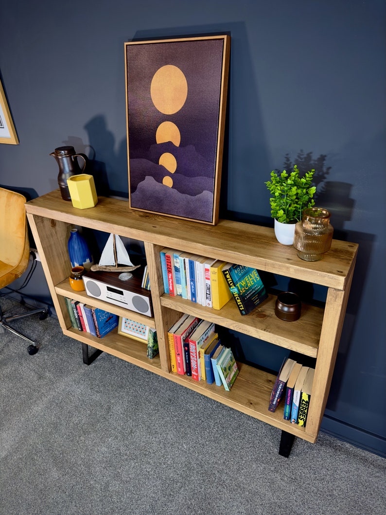 May include: A wooden bookshelf with black metal legs, filled with books, decorative items, and a framed artwork depicting golden orbs over a purple landscape. A small white radio and a miniature sailboat are also visible.