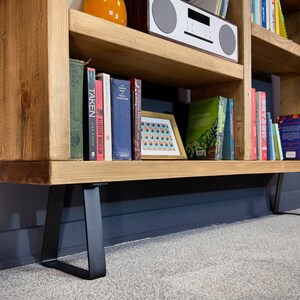 May include: A light-colored wooden bookshelf with black metal legs. The shelves are filled with books of various sizes and colors, a framed picture, and a white radio. The bookshelf is against a dark gray wall.