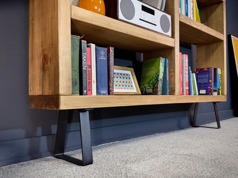May include: A light-colored wooden bookshelf with black metal legs. The shelves are filled with books of various sizes and colors, a framed picture, and a white radio. The bookshelf is against a dark gray wall.
