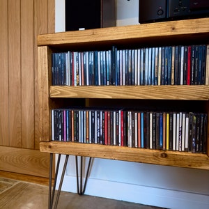 May include: A rustic wooden CD storage cabinet with two shelves, filled with compact discs. The cabinet has a warm, natural wood finish and is supported by hairpin legs. A speaker and stereo equipment sit on top.