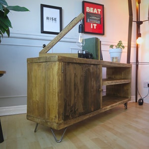 K&ouml;nnte beinhalten: Rustikaler TV-Schrank aus Holz mit Schrank und offenen Regalen. Der Schrank hat eine nat&uuml;rliche Holzoberfl&auml;che, Haarnadelbeine aus Metall und eine eingebaute Lampe. Ein gerahmter Druck mit dem Wort "RISE" befindet sich an der Wand.