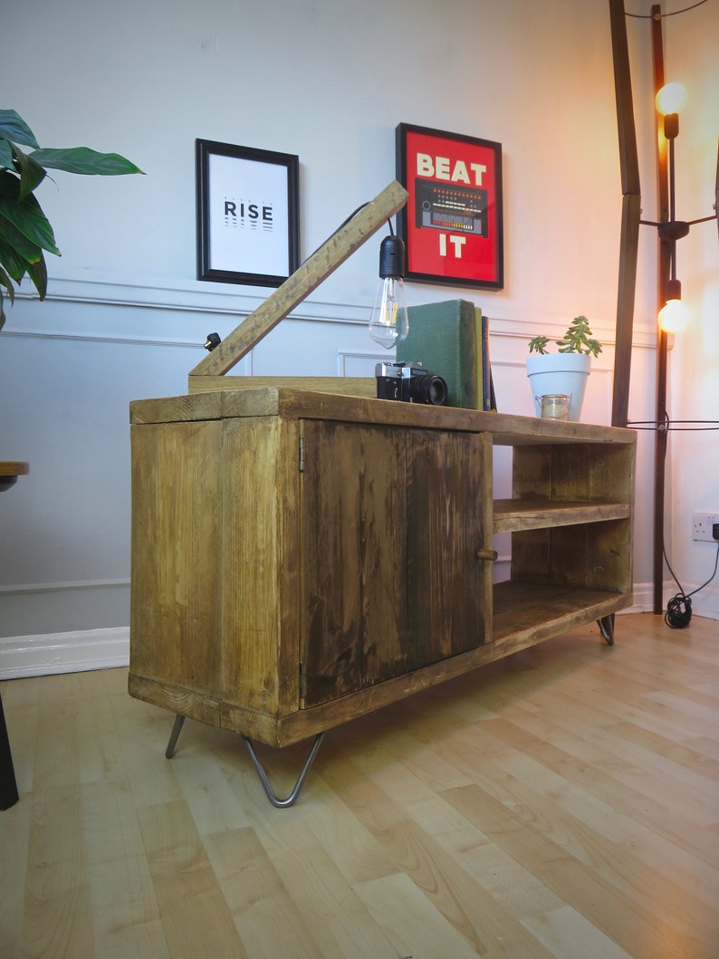 K&ouml;nnte beinhalten: Rustikaler TV-Schrank aus Holz mit Schrank und offenen Regalen. Der Schrank hat eine nat&uuml;rliche Holzoberfl&auml;che, Haarnadelbeine aus Metall und eine eingebaute Lampe. Ein gerahmter Druck mit dem Wort "RISE" befindet sich an der Wand.