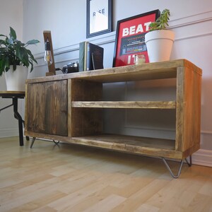 K&ouml;nnte beinhalten: Rustikale TV-Konsole aus Holz mit einer dunkel gebeizten T&uuml;r und zwei offenen Regalen. Die Konsole hat Haarnadelbeine und ist mit Dekorationen wie einem gerahmten Poster mit der Aufschrift "BEAT IT" versehen. Eine Topfpflanze steht oben drauf. Die Konsole ist 120 cm breit.