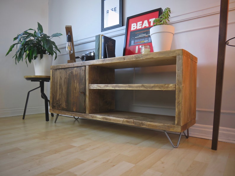 K&ouml;nnte beinhalten: Rustikale TV-Konsole aus Holz mit einer dunkel gebeizten T&uuml;r und zwei offenen Regalen. Die Konsole hat Haarnadelbeine und ist mit Dekorationen wie einem gerahmten Poster mit der Aufschrift "BEAT IT" versehen. Eine Topfpflanze steht oben drauf. Die Konsole ist 120 cm breit.