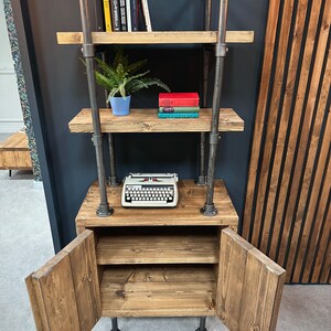 以下が含まれることがあります： 明るい木材と暗い金属パイプで作られた、インダストリアルスタイルの本棚。オープンシェルフとキャビネットがあり、ヴィンテージのタイプライターと本が飾られています。小さな鉢植えの植物が置かれています。木材は明るい茶色です。