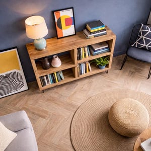 Op de afbeelding: Een gezellige woonkamer met een houten boekenkast, een rond jute vloerkleed en een lichtblauwe lamp. Kunstwerken en planten voegen decoratieve accenten toe. Een grijze stoel met een geometrisch kussen is zichtbaar.
