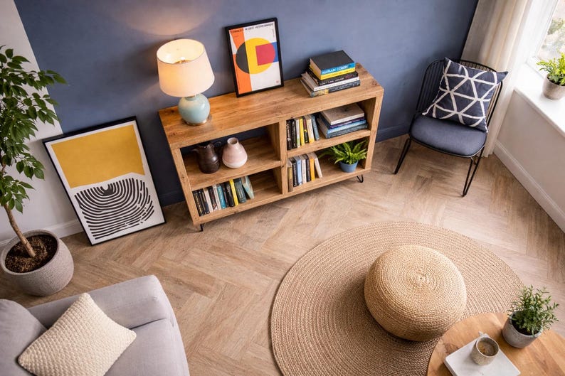 Op de afbeelding: Een gezellige woonkamer met een houten boekenkast, een rond jute vloerkleed en een lichtblauwe lamp. Kunstwerken en planten voegen decoratieve accenten toe. Een grijze stoel met een geometrisch kussen is zichtbaar.