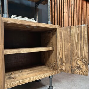 以下が含まれることがあります： 扉が開いた素朴な木製キャビネット。内部の棚が見えます。キャビネットは着色された木材で構成され、金属パイプで支えられています。キャビネットの上にある棚には、ヴィンテージのタイプライターが置かれています。
