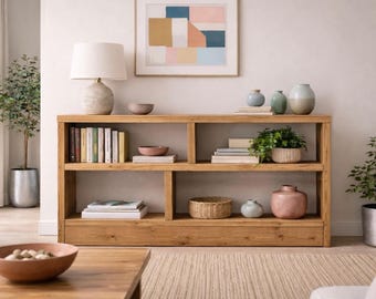 Könnte beinhalten: Ein hellbraunes Bücherregal aus Holz mit mehreren Regalen, das Bücher, Pflanzen und dekorative Gegenstände enthält. Die Regale sind mit Büchern, kleinen Vasen und einem geflochtenen Korb gefüllt. Eine Lampe und ein Kunstwerk sind ebenfalls zu sehen.