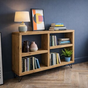 Op de afbeelding: Een houten boekenplank met meerdere compartimenten, gevuld met boeken en decoratieve items. Een lamp met een beige kap staat bovenop, naast een ingelijste abstracte kunstprint. De boekenplank wordt ondersteund door zwarte metalen poten.