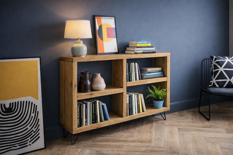 Op de afbeelding: Een houten boekenplank met meerdere compartimenten, gevuld met boeken en decoratieve items. Een lamp met een beige kap staat bovenop, naast een ingelijste abstracte kunstprint. De boekenplank wordt ondersteund door zwarte metalen poten.