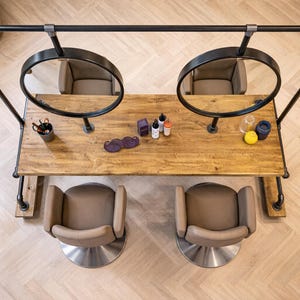 K&ouml;nnte beinhalten: Draufsicht auf eine Salonstation mit zwei runden Spiegeln, zwei St&uuml;hlen und einem Holztisch. Auf dem Tisch befinden sich verschiedene Sch&ouml;nheitsprodukte und ein schnurrbartf&ouml;rmiges Accessoire. Die Station ist aus schwarzen Metallrohren und -fittings gefertigt.
