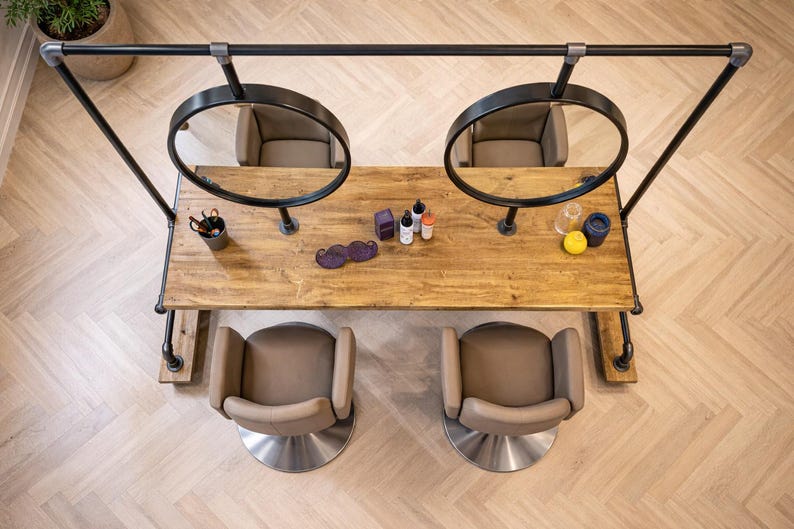 K&ouml;nnte beinhalten: Draufsicht auf eine Salonstation mit zwei runden Spiegeln, zwei St&uuml;hlen und einem Holztisch. Auf dem Tisch befinden sich verschiedene Sch&ouml;nheitsprodukte und ein schnurrbartf&ouml;rmiges Accessoire. Die Station ist aus schwarzen Metallrohren und -fittings gefertigt.