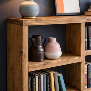 Op de afbeelding: Houten boekenplank met drie planken, met boeken, een blauwe keramische lamp en decoratieve vazen. De boekenplank is gemaakt van lichtbruin hout en heeft een eenvoudig, modern ontwerp. Bovenop staat een ingelijste abstracte kunstprint.
