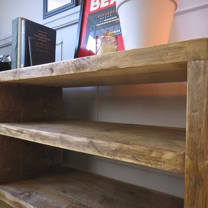 K&ouml;nnte beinhalten: Rustikales TV-M&ouml;bel aus Holz mit zwei offenen Regalen und einer Schrankt&uuml;r. Das Holz hat einen warmen, braunen Farbton mit sichtbarer Maserung. Auf dem oberen Regal befinden sich B&uuml;cher, eine Kamera und eine dekorative Lampe.