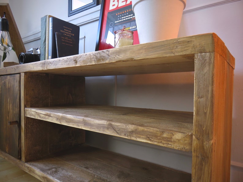 K&ouml;nnte beinhalten: Rustikales TV-M&ouml;bel aus Holz mit zwei offenen Regalen und einer Schrankt&uuml;r. Das Holz hat einen warmen, braunen Farbton mit sichtbarer Maserung. Auf dem oberen Regal befinden sich B&uuml;cher, eine Kamera und eine dekorative Lampe.