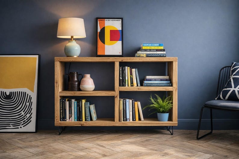 Op de afbeelding: Houten boekenkast met meerdere compartimenten, gevuld met boeken, decoratieve vazen en een potplant. Een tafellamp met een beige kap en een ingelijste abstracte kunstprint staan erop. De kamer heeft een blauwe muur en een houten vloer.