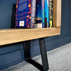May include: A wooden bookshelf with black metal legs. Several books with colorful spines are displayed on the shelf. The books have titles such as "Museums & Broken Promises" and "Zeroes". The bookshelf is set against a dark blue wall and a gray carpet.