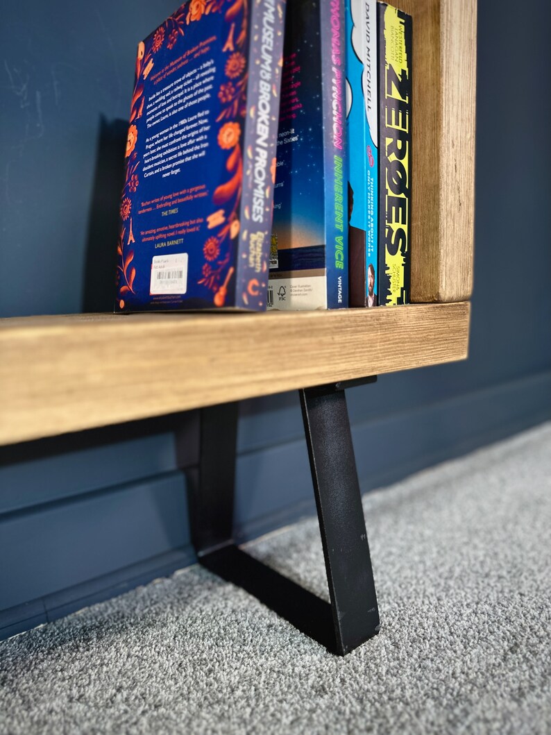 May include: A wooden bookshelf with black metal legs. Several books with colorful spines are displayed on the shelf. The books have titles such as "Museums & Broken Promises" and "Zeroes". The bookshelf is set against a dark blue wall and a gray carpet.