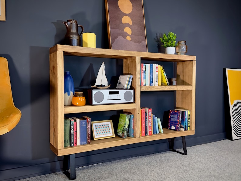 May include: A wooden bookshelf with multiple shelves, displaying books, decorative items, and a small radio. The bookshelf has a natural wood finish and black metal legs. Various decorative items are placed on top of the bookshelf.