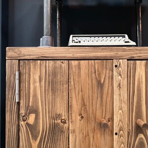 以下が含まれることがあります： 自然な仕上がりの素朴な木製キャビネット。3つのドアと木製のノブが付いています。上にはヴィンテージのタイプライターがあり、背景には金属パイプと濃い青色の壁があります。キャビネットの木目は際立っています。