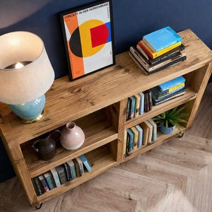 Op de afbeelding: Houten boekenkast met meerdere planken gevuld met boeken, een kleine plant in een pot en decoratieve vazen. Een lamp met een blauwe voet en een ingelijste kunstdruk staan erop. De boekenkast is gemaakt van lichtgekleurd hout.