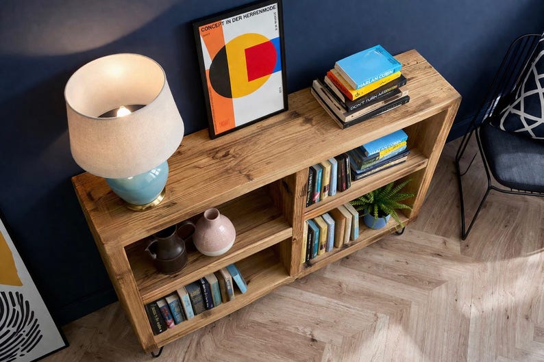 Op de afbeelding: Houten boekenkast met meerdere planken gevuld met boeken, een kleine plant in een pot en decoratieve vazen. Een lamp met een blauwe voet en een ingelijste kunstdruk staan erop. De boekenkast is gemaakt van lichtgekleurd hout.