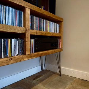 May include: Wooden media console with multiple shelves filled with CDs. The console has a natural wood finish and is supported by hairpin legs. A black audio receiver is visible in the center shelf.