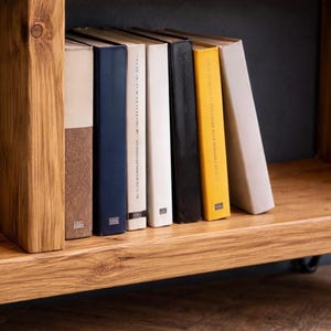Op de afbeelding: Een houten boekenplank met een verzameling boeken. De boeken hebben verschillende gekleurde ruggen, waaronder blauw, bruin, wit, zwart en geel. De plank is gemaakt van lichtgekleurd hout.
