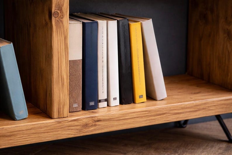 Op de afbeelding: Een houten boekenplank met een verzameling boeken. De boeken hebben verschillende gekleurde ruggen, waaronder blauw, bruin, wit, zwart en geel. De plank is gemaakt van lichtgekleurd hout.