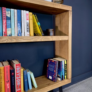 May include: A wooden bookshelf with a natural finish, featuring multiple shelves filled with books of various sizes and colors. The bookshelf has a modern design with black metal legs and is set against a dark blue wall.