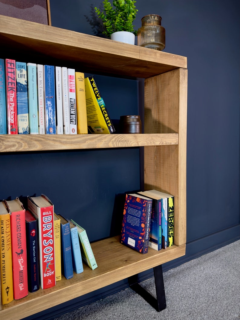 May include: A wooden bookshelf with a natural finish, featuring multiple shelves filled with books of various sizes and colors. The bookshelf has a modern design with black metal legs and is set against a dark blue wall.