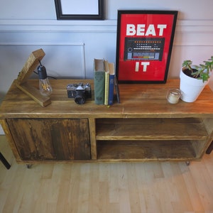 K&ouml;nnte beinhalten: Ein rustikaler TV-Schrank aus Holz mit einer Schiebet&uuml;r und offenen Regalen. Der Schrank ist mit einer Vintage-Kamera, B&uuml;chern und einem gerahmten Poster mit der Aufschrift "BEAT IT" versehen. Eine Holzlampe und eine Topfpflanze erg&auml;nzen die Einrichtung.