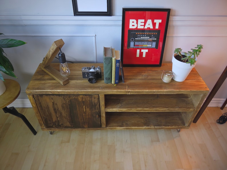 K&ouml;nnte beinhalten: Ein rustikaler TV-Schrank aus Holz mit einer Schiebet&uuml;r und offenen Regalen. Der Schrank ist mit einer Vintage-Kamera, B&uuml;chern und einem gerahmten Poster mit der Aufschrift "BEAT IT" versehen. Eine Holzlampe und eine Topfpflanze erg&auml;nzen die Einrichtung.