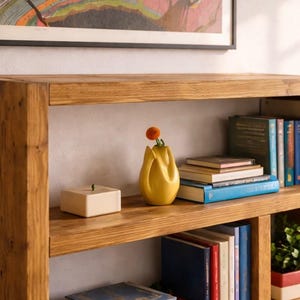 Puede incluir: Una estanter&iacute;a de madera con tres estantes, llena de libros, un jarr&oacute;n amarillo con una flor y una peque&ntilde;a caja blanca. La estanter&iacute;a es de madera marr&oacute;n claro y est&aacute; contra una pared blanca.