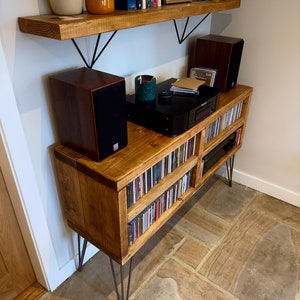 May include: A wooden media console with hairpin legs, filled with CDs. Two speakers sit on top, along with a CD player and decorative items. A wooden shelf above holds books and decor. The console is made of light brown wood.