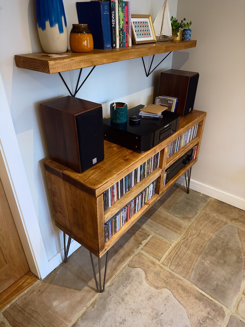 May include: A wooden media console with hairpin legs, filled with CDs. Two speakers sit on top, along with a CD player and decorative items. A wooden shelf above holds books and decor. The console is made of light brown wood.