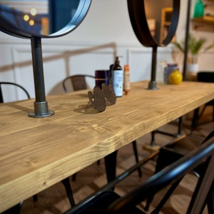 K&ouml;nnte beinhalten: Ein langer, hellbrauner Holztisch mit schwarzen Metallst&uuml;hlen und zwei runden Spiegeln. Der Tisch hat eine nat&uuml;rliche Holzmaserung und ist von schwarzen Metallst&uuml;hlen umgeben. Auf dem Tisch befinden sich verschiedene Flaschen und ein dekoratives Metallobjekt.