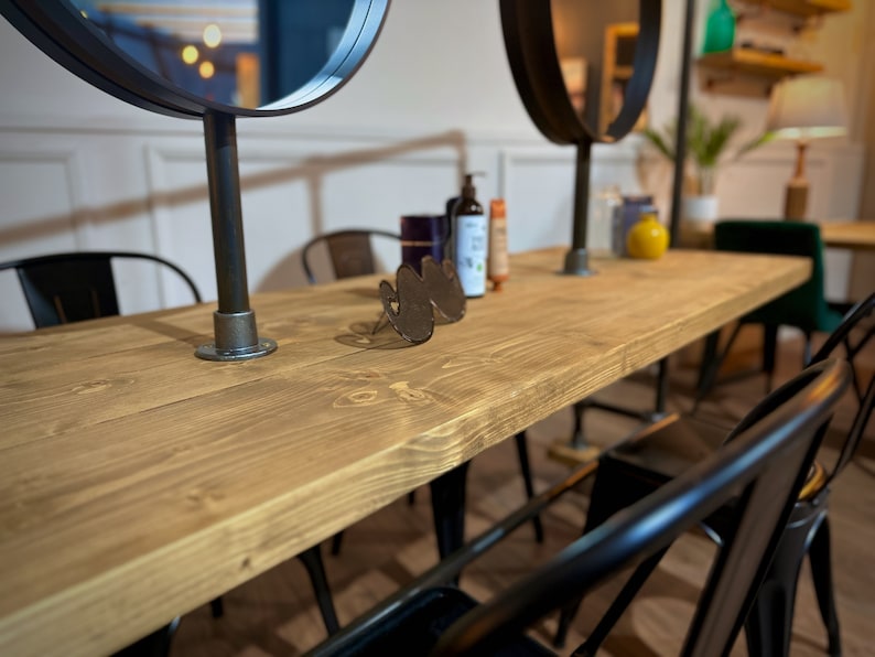 K&ouml;nnte beinhalten: Ein langer, hellbrauner Holztisch mit schwarzen Metallst&uuml;hlen und zwei runden Spiegeln. Der Tisch hat eine nat&uuml;rliche Holzmaserung und ist von schwarzen Metallst&uuml;hlen umgeben. Auf dem Tisch befinden sich verschiedene Flaschen und ein dekoratives Metallobjekt.