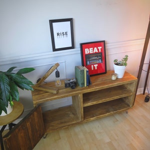 K&ouml;nnte beinhalten: Rustikale TV-Konsole aus Holz mit offenen Regalen und einer Schrankt&uuml;r. Die Konsole zeigt ein gerahmtes Poster mit der Aufschrift "BEAT IT" und ein weiteres mit der Aufschrift "RISE". Eine Vintage-Kamera, B&uuml;cher und eine kleine Pflanze stehen obenauf.