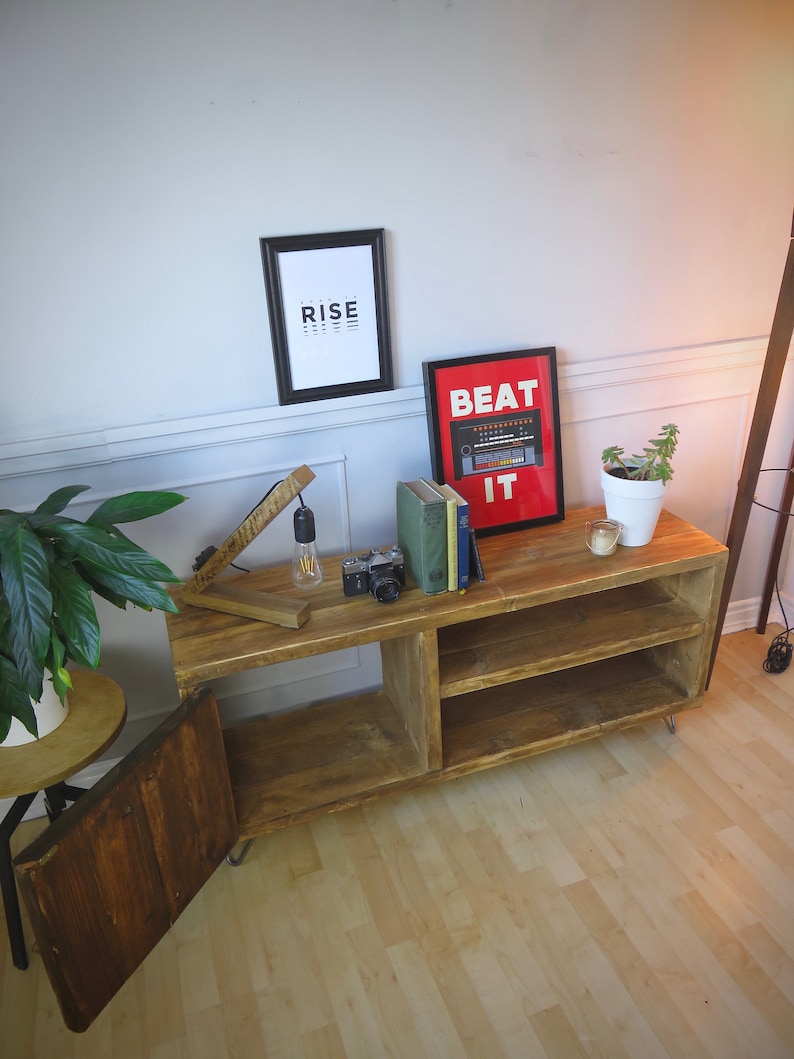 K&ouml;nnte beinhalten: Rustikale TV-Konsole aus Holz mit offenen Regalen und einer Schrankt&uuml;r. Die Konsole zeigt ein gerahmtes Poster mit der Aufschrift "BEAT IT" und ein weiteres mit der Aufschrift "RISE". Eine Vintage-Kamera, B&uuml;cher und eine kleine Pflanze stehen obenauf.