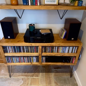 May include: A wooden media console with a natural finish, featuring a black stereo receiver, two dark brown speakers, and storage for CDs. The console is supported by metal hairpin legs and has a matching wooden shelf above.