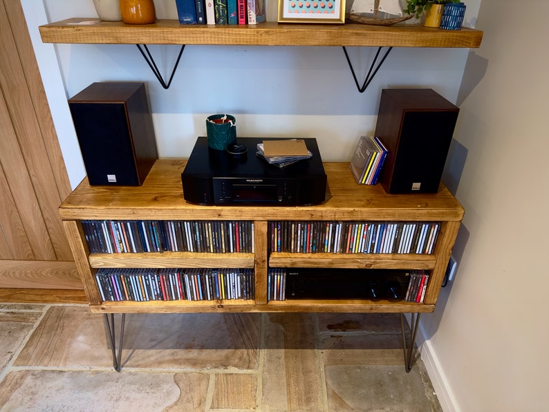 May include: A wooden media console with a natural finish, featuring a black stereo receiver, two dark brown speakers, and storage for CDs. The console is supported by metal hairpin legs and has a matching wooden shelf above.